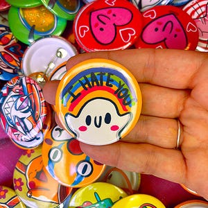 Peut inclure: Un badge rond avec une épingle représentant un personnage blanc ressemblant à un fantôme avec des joues roses et un arc-en-ciel au-dessus. Le texte I HATE YOU est écrit en lettres noires sur l'arc-en-ciel. Le badge a une bordure orange vif.