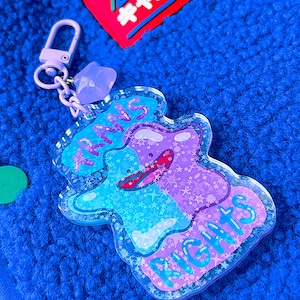 Può includere: Un portachiavi in acrilico raffigurante un personaggio a forma di blob diviso in blu e viola con una finitura olografica a motivi stellati. Il testo TRANS RIGHTS è stampato in blu e viola. Include una chiusura viola chiaro e un ciondolo a stella.