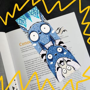 Peut inclure: Un marque-page bleu avec une illustration de dessin animé de deux créatures aux grands yeux et aux oreilles pointues. Le marque-page a un fond bleu clair avec un motif de feuilles en haut. Le marque-page est posé sur un livre ouvert.