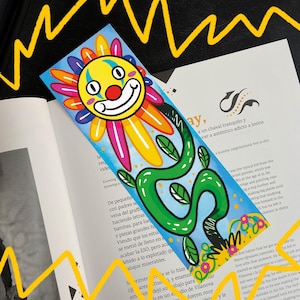 Peut inclure: Un marque-page coloré avec un visage de clown souriant et un motif floral. Le marque-page a un fond bleu avec une vigne et des feuilles vertes. Le marque-page est placé sur un livre ouvert.