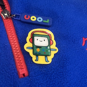 Peut inclure: Veste polaire bleue avec une fermeture éclair rouge et une tirette de fermeture éclair bleue avec le mot "cool" en lettres colorées. Un autocollant jaune d'un personnage de dessin animé avec un casque est également sur la veste.