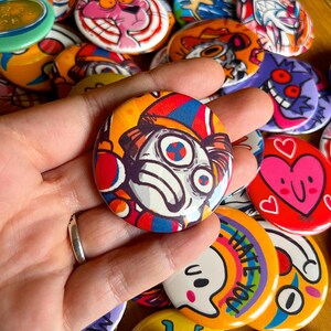 Könnte beinhalten: Eine Sammlung bunter, runder Ansteckbuttons mit verschiedenen Cartoon-Designs. Ein Button zeigt einen clownartigen Charakter mit rotem und blauem Farbschema. Andere Buttons zeigen einen rosa Panther, einen Geist und ein Herz mit einem Gesicht.