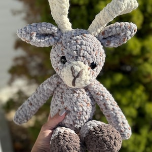 May include: A crocheted plush deer toy with brown and white antlers, a gray and brown speckled body, and brown feet.