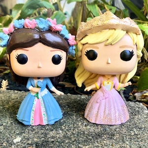May include: Two Funko Pop figures of princesses. One figure wears a blue dress with a pink underskirt and a floral crown. The other figure wears a pink dress with a gold crown and blonde hair. Both figures have large black eyes.