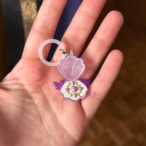 Puede incluir: Un llavero en forma de concha de color lavanda con un anillo transparente. La concha se abre para revelar un diseño blanco y dorado con detalles morados. El llavero está hecho de plástico y tiene un pequeño cierre de metal.
