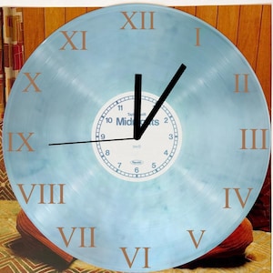 Puede incluir: Un reloj de vinilo azul claro con marcadores de hora en números romanos. La esfera del reloj tiene un centro blanco con manecillas negras y una esfera de reloj blanca más pequeña con números negros del 1 al 12. El texto "Taylor Swift Midnights" está impreso en la esfera más pequeña.