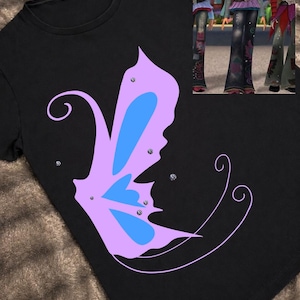 May include: Black t-shirt featuring a large butterfly design in light purple and blue, embellished with small rhinestones. The butterfly has a stylized wing shape. The shirt is a casual wear item.