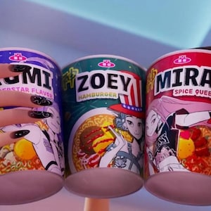 Könnte beinhalten: Drei bunte Ramen-Nudeltassen werden gezeigt. Die Tassen sind mit "KUMI", "ZOEY" und "MIRA" beschriftet. Jede Tasse zeigt eine einzigartige Illustration und Geschmacksbeschreibung. Die "ZOEY"-Tasse hat eine Hamburger-Grafik.