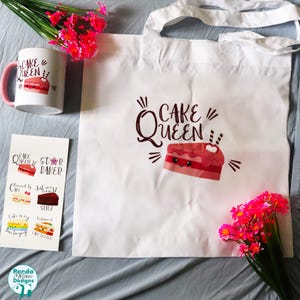 May include: A white tote bag and a white ceramic mug with a pink handle, both featuring a graphic of a smiling pink cake slice and the text "CAKE QUEEN". A rectangular sticker sheet displays six cake-themed illustrations and text labels.