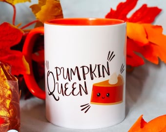 May include: A white ceramic mug with an orange interior and handle. The front features the text PUMPKIN QUEEN in dark brown lettering next to a graphic of a smiling slice of pumpkin pie with a dollop of whipped cream on top.