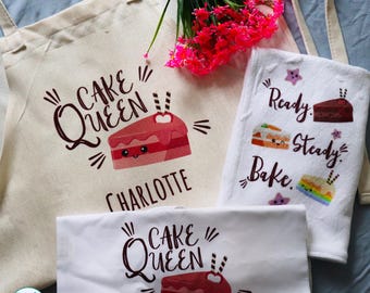 May include: A beige canvas apron, a white towel, and a white folded fabric piece, each featuring a graphic of a smiling cake slice and the text "Cake Queen" or "Ready. Steady. Bake." The name "Charlotte" is printed on the apron.