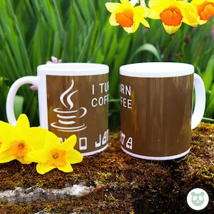 May include: Two brown ceramic coffee mugs with white text and a java logo. The mug on the left says "I turn coffee into java". The mug on the right says "I turn coffee into java". The mugs are sitting on a bed of green moss with yellow daffodils.
