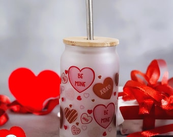 May include: A frosted glass can-shaped tumbler with a bamboo lid and a metal straw. The exterior features a pattern of red, pink, and brown hearts with the text BE MINE, XOXO, and HUGS printed in red and brown on the surface of the glass.