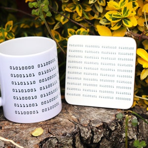 May include: A white ceramic mug and square coaster set, both featuring a black binary code design. The mug has a curved handle. The items are displayed on a wooden surface, with green and yellow foliage in the background. The coaster is square.