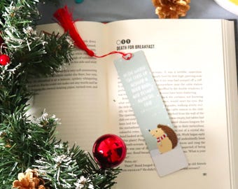 May include: Open book with a red tassel bookmark and a decorative bookmark featuring a hedgehog illustration. The bookmark has text that reads "In the middle of the chaos of a good book, are you taking a moment to breathe?"