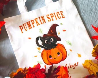 May include: A white tote bag featuring a graphic of a black cat wearing a brown witch hat sitting on an orange pumpkin. The text PUMPKIN SPICE is printed in orange above the cat. Small colorful leaves are scattered around the central illustration.