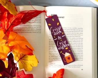 May include: A bookmark with the text "Crisp days & Cozy nights" and a graphic of a mug with whipped cream. The bookmark is purple with yellow leaf accents and a red tassel. Autumn leaves surround an open book.