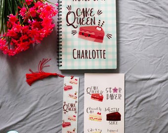 May include: A spiral-bound notebook with a light blue checkered cover featuring the text Notes Of A Cake Queen and Charlotte, a matching bookmark with a red tassel, and a sticker sheet with various cake illustrations and text labels.