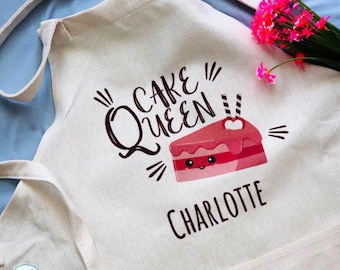 May include: A beige apron featuring the text "CAKE QUEEN" in dark brown, a graphic of a pink cake slice with a smiling face, and the name "CHARLOTTE" printed below. A small bouquet of bright pink flowers is tucked into the top corner of the apron.