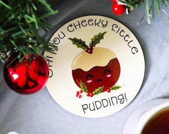 May include: A round coaster featuring a cartoon Christmas pudding with a smiling face, topped with holly leaves and berries. The text reads, Oh you cheeky little pudding! The design includes a small candy cane and is printed on a cream background.