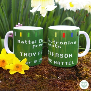 May include: Two green ceramic mugs with white handles. The mugs feature a pixelated design with the text "Mattel Electronics Presents Troy Peterson 82 Mattel" in white.