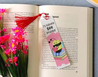 May include: A colorful bookmark with a red tassel is placed in an open book. The bookmark features the text "I'D RATHER BEE READING" and an illustration of a bee reading a book. Pink flowers are in the foreground.