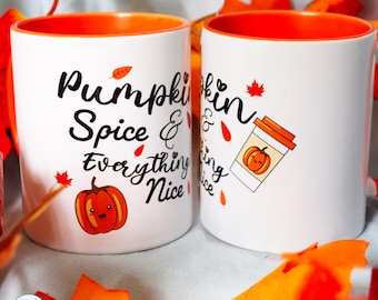 May include: Two white ceramic mugs with bright orange interiors. The mugs feature black cursive text reading Pumpkin Spice & Everything Nice, decorated with small orange pumpkin illustrations with faces and scattered red and orange autumn leaves.