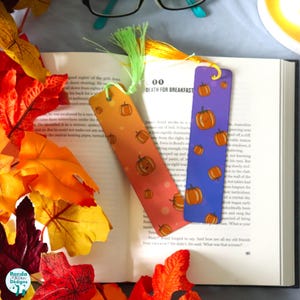 May include: Two decorative bookmarks with pumpkin designs. One bookmark has an orange gradient background, and the other has a purple gradient. Both have a green tassel. The bookmarks are on top of an open book with fall leaves.