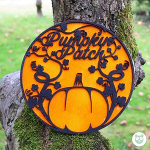 May include: A round wooden sign with a pumpkin patch theme. The sign is painted orange and features a black silhouette of a pumpkin with vines and leaves. The text "Pumpkin Patch" is written in black on the sign.