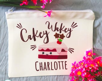 May include: A white zippered pouch featuring the dark brown text "Cakey Wakey" above a graphic of a smiling pink and red cake slice topped with a strawberry and two star-shaped decorations, with the name "CHARLOTTE" printed in dark brown below.