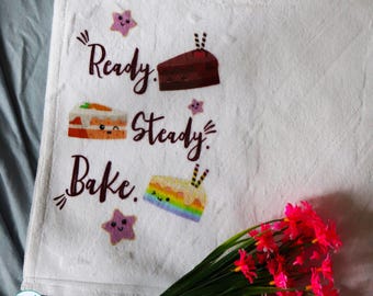 May include: A white kitchen towel featuring the text Ready, Steady, Bake in dark brown script. The design includes three illustrated cake slices with cute faces, including a chocolate slice, a carrot cake slice, and a rainbow cake slice.