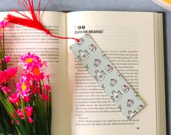 May include: A bookmark with a light blue background featuring a pattern of aprons and teacups, resting on an open book. The book has the title "DEATH FOR BREAKFAST" and is surrounded by pink flowers and a red tassel.