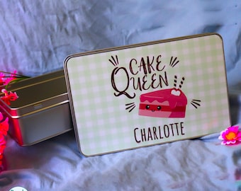 May include: A rectangular metal tin with a light green gingham pattern on the lid. The lid features the text CAKE QUEEN above a graphic of a pink cake slice with a smiling face and the name CHARLOTTE printed in black lettering below the cake.