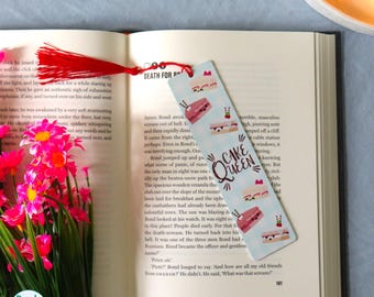 May include: A bookmark with a red tassel rests on an open book. The bookmark features a light blue background with cake illustrations and the text "Cake Queen". Pink flowers are in the foreground.