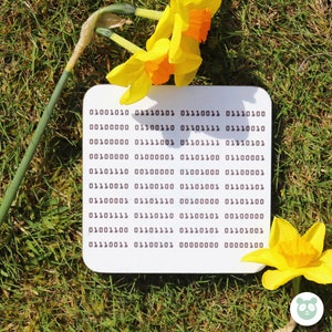 May include: A white card with a pattern of 0s and 1s, resembling binary code. The card is lying on a bed of green grass with yellow daffodils.