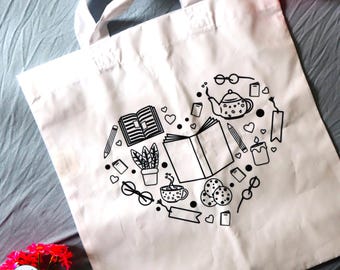 May include: A white tote bag featuring a black line art graphic arranged in a heart shape. The design includes illustrations of open books, a teapot, a potted plant, a steaming mug, cookies, eyeglasses, pencils, candles, and small hearts.