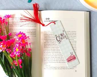 May include: A bookmark with a red tassel, featuring the text "Cake Queen" and a cake illustration, rests on an open book. Pink flowers and eyeglasses are also visible, creating a cozy reading scene.