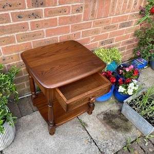 May include: A wooden side table with a drawer, in a warm brown finish. The table has a square top, a lower shelf, and turned legs. The drawer is open, revealing the interior. The table is approximately 61 cm tall.