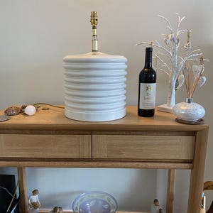May include: A white, ribbed ceramic lamp stands on a wooden table. A bottle of red wine and decorative items are also present. The lamp features a brass-coloured neck and a light bulb socket.
