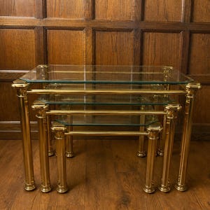 May include: A set of three nesting tables featuring rectangular clear glass tops and polished gold-tone metal frames. Each table has four vertical cylindrical legs with decorative caps at the top and bottom, creating a tiered arrangement.