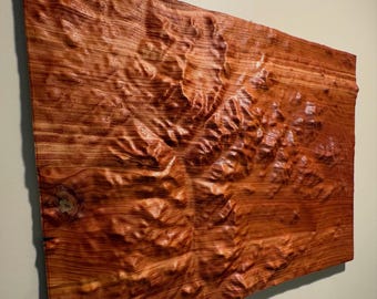 Könnte beinhalten: Ein rechteckiges Wandkunstobjekt aus Holz mit einer geschnitzten topografischen Reliefkarte. Die Oberfläche zeigt eine satte rotbraune Holzmaserung mit sichtbaren Ästen und tiefen, strukturierten Konturen, die Gebirgszüge nachbilden.