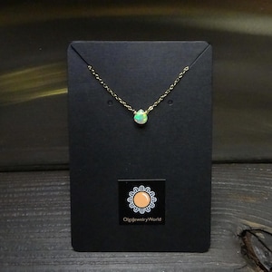Può includere: Una delicata collana d'oro con un piccolo pendente di opale a forma di goccia. L'opale mostra un mix di colori, tra cui bianco, verde e sfumature di blu. La collana è esposta su una carta nera con il logo "OlgajewelryWorld".
