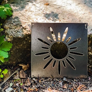 Op de afbeelding: Een metalen zonuitsparing met een rustieke afwerking. De zon heeft een cirkelvormig midden en 12 stralen die naar buiten uitstrekken.
