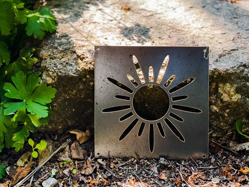 Op de afbeelding: Een metalen zonuitsparing met een rustieke afwerking. De zon heeft een cirkelvormig midden en 12 stralen die naar buiten uitstrekken.