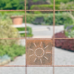 Pu&ograve; includere: Un designo di sole in metallo arrugginito tagliato da una lamiera di metallo quadrata. Il sole &egrave; delineato in bianco e ha raggi che si estendono dal cerchio.