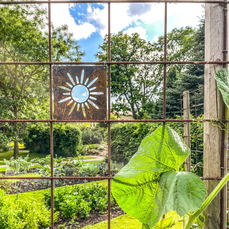 Op de afbeelding: Een roestige metalen zon-uitsparing is bevestigd aan een draadhek. De zon bevindt zich in het midden van de afbeelding en is omgeven door een groene tuin.