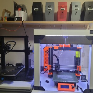 May include: Two 3D printers, one black and one white, with a clear enclosure. The white printer has an orange frame and a glass build plate. The black printer has a black build plate. Both printers are on a shelf with several black, red, and grey boxes.
