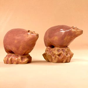May include: Two ceramic bear figurines in a reddish-brown glaze. The bears are perched on textured, rock-like bases. One bear is in a seated position, while the other is standing. The background is a solid, light peach color.