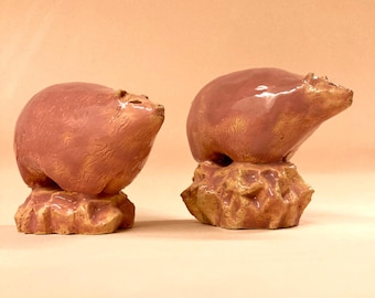 May include: Two ceramic bear figurines in a reddish-brown glaze. The bears are perched on textured, rock-like bases. One bear is in a seated position, while the other is standing. The background is a solid, light peach color.