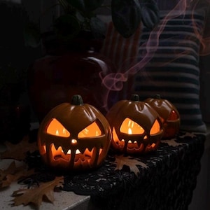 Può includere: Tre lanterne di ceramica arancione a forma di zucca con facce intagliate e interni luminosi. Le zucche sono su un panno di pizzo nero con foglie autunnali. Uno sfondo scuro include un vaso e un cuscino.